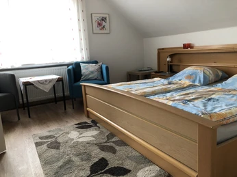 Schlafbereich Helles Doppelbettzimmer mit blau-grauer Bettwäsche, zwei Sesseln, einem Tisch und einem Teppich.