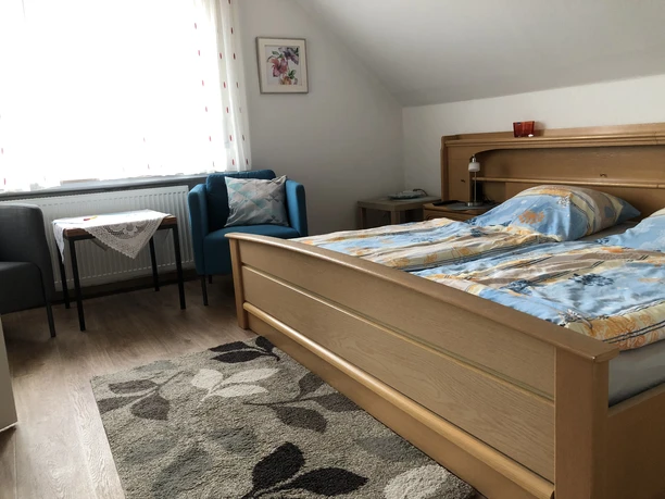 Schlafbereich Helles Doppelbettzimmer mit blau-grauer Bettwäsche, zwei Sesseln, einem Tisch und einem Teppich.