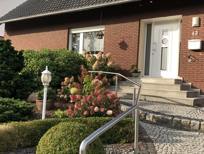 Eingangsbereich Eingangsbereich eines roten Backsteinhauses mit Blumenbeeten, Treppe und Handlauf, ergänzt durch Laterne.