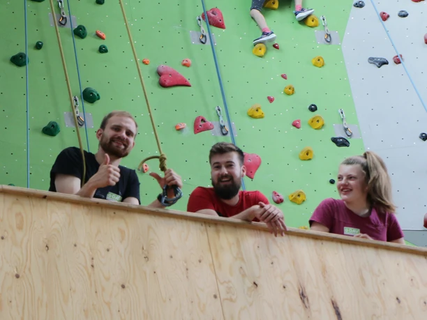 Drei junge Erwachsene lächeln und zeigen Daumen hoch vor einer bunten Kletterwand.