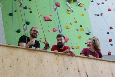 Kletterzentrum Drei junge Erwachsene lächeln und zeigen Daumen hoch vor einer bunten Kletterwand.