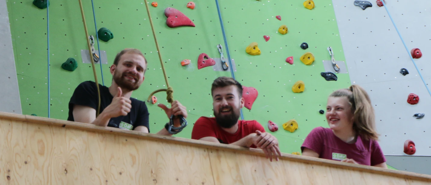 Kletterzentrum Drei junge Erwachsene lächeln und zeigen Daumen hoch vor einer bunten Kletterwand.