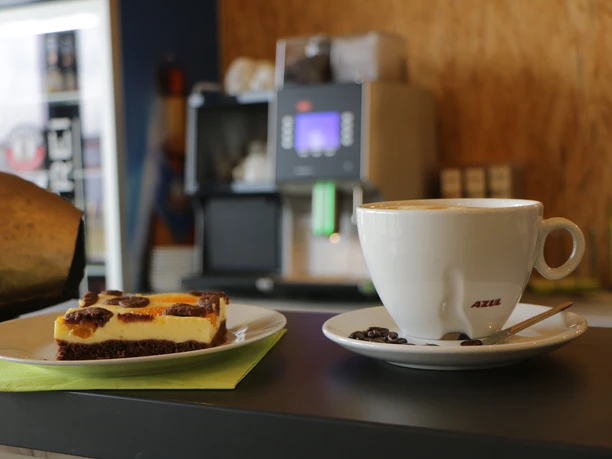 KLZ Kaffee Kuchen.JPG Kaffee auf Untertasse neben einem Stück Kuchen auf grünem Serviettenpapier, im Café serviert.
