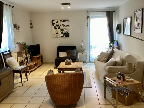 Fewo Lustig_GL.jpeg Gemütliches Wohnzimmer mit beige Sofa, Rattansessel, Wanddekorationen und großen Fenstern für viel Licht.