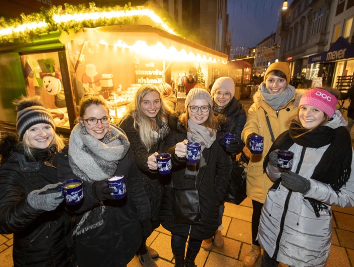 Eine Gruppe von sieben Personen hält Tassen mit Glühwein, fröhlich vor einem Weihnachtsmarktstand.