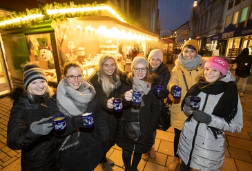 Eine Gruppe von sieben Personen hält Tassen mit Glühwein, fröhlich vor einem Weihnachtsmarktstand.