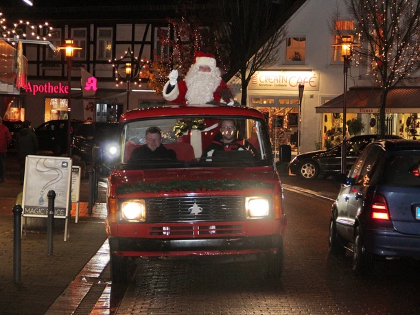 Weihnachtsmann winkt von einem roten Oldtimer im festlich beleuchteten Stadtzentrum aus.