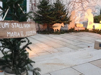 Weihnachtliche Krippenszene vor einer Mauer, beleuchtet, mit Schildern und Tannenästen dekoriert.