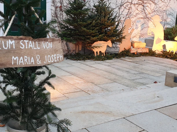 Weihnachtliche Krippenszene vor einer Mauer, beleuchtet, mit Schildern und Tannenästen dekoriert.