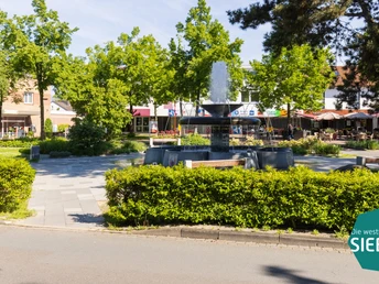 Brunnen auf einem Platz mit umliegenden Bänken und Bäumen in einer sonnigen Geschäftsumgebung.
