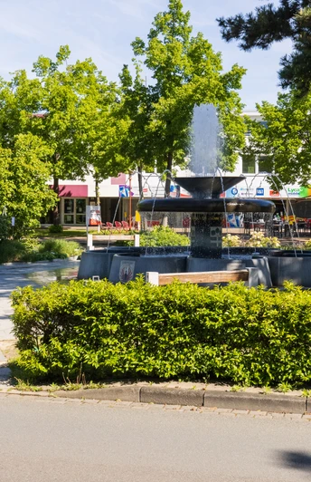 Brunnen auf einem Platz mit umliegenden Bänken und Bäumen in einer sonnigen Geschäftsumgebung.