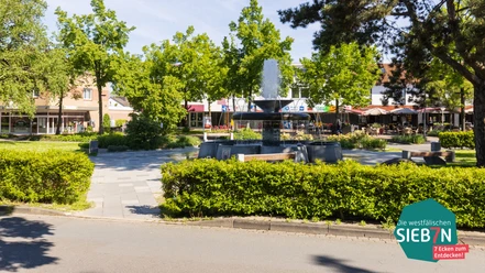 Espelkamp Brunnen auf einem Platz mit umliegenden Bänken und Bäumen in einer sonnigen Geschäftsumgebung.