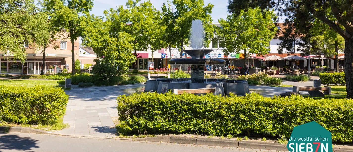 Espelkamp Brunnen auf einem Platz mit umliegenden Bänken und Bäumen in einer sonnigen Geschäftsumgebung.