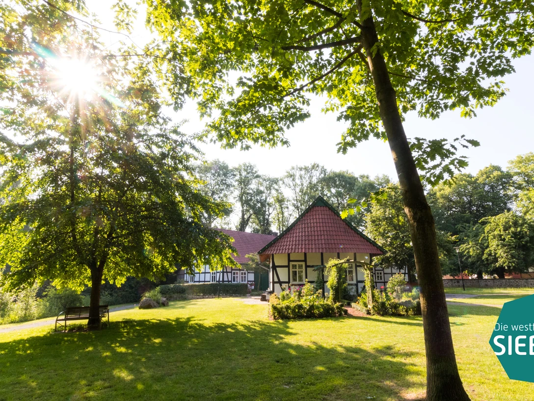 Hille Traditionelles Fachwerkhaus im dichten grünen Wald, direkte Sonne im Hintergrund, ruhig gelegen.