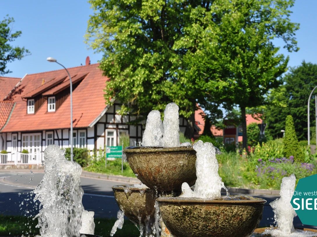 Huellhorst.JPG Ein dreistufiger Brunnen vor Fachwerkhäusern und üppigem Grün an einer sonnigen Kreuzung in Hüllhorst.