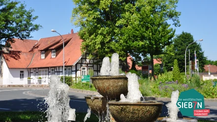 Huellhorst Ein dreistufiger Brunnen vor Fachwerkhäusern und üppigem Grün an einer sonnigen Kreuzung in Hüllhorst.