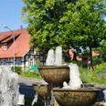 Huellhorst Ein dreistufiger Brunnen vor Fachwerkhäusern und üppigem Grün an einer sonnigen Kreuzung in Hüllhorst.