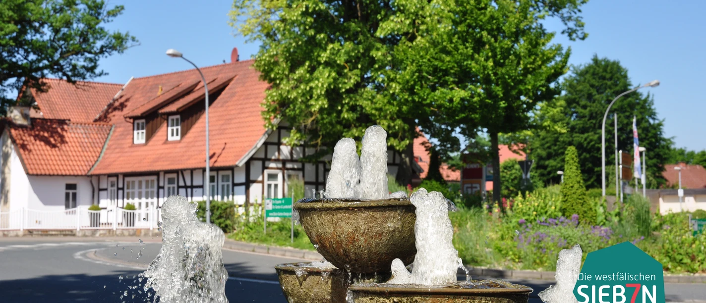Huellhorst Ein dreistufiger Brunnen vor Fachwerkhäusern und üppigem Grün an einer sonnigen Kreuzung in Hüllhorst.