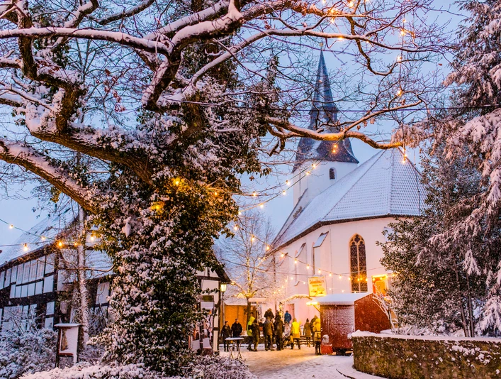 Stemwede-Levern Weihnachten Verschneiter Kirchenplatz in Stemwede-Levern, umgeben von Fachwerkhäusern, Lichterketten funkeln.
