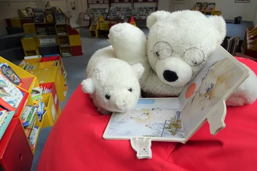 Zwei Plüsch-Eisbären lesen gemeinsam ein Kinderbuch in der bunten Leseecke einer Bibliothek.