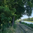 Gastro Preußisch Oldendorf Zwei Menschen spazieren auf einem schattigen Weg entlang eines Kanals, umgeben von Bäumen.
