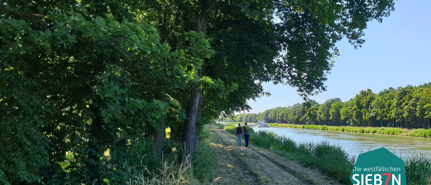 Gastro Preußisch Oldendorf Zwei Menschen spazieren auf einem schattigen Weg entlang eines Kanals, umgeben von Bäumen.