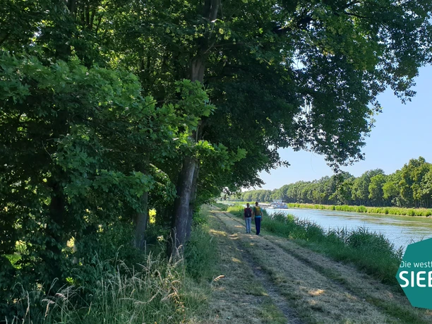 Gastro Preußisch Oldendorf Zwei Menschen spazieren auf einem schattigen Weg entlang eines Kanals, umgeben von Bäumen.