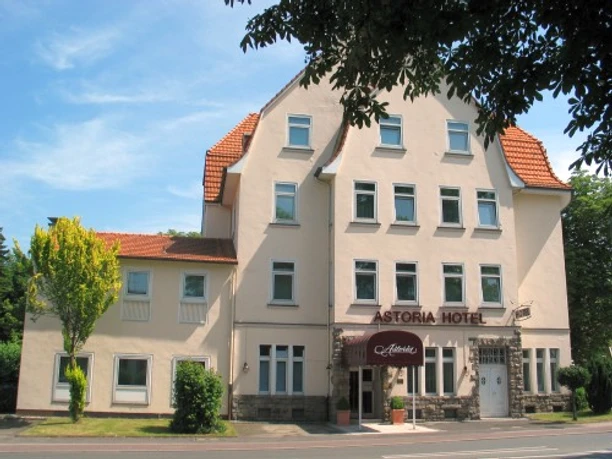 Hotel ASTORIA Ratingen Drei-stöckiges Gebäude mit rotem Ziegeldach im Grünen, beiger Fassade und Hotelbeschilderung vorne.