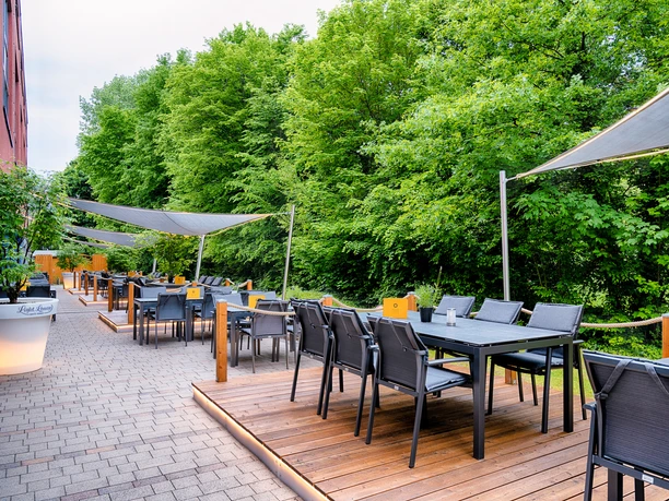 Welcome Hotel Paderborn / Light Lounge Bar & Terrace Sonnige Terrasse mit modernen Sitzmöbeln im Schatten von Markisen und umgeben von grünem Laubwerk.