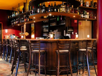 Welcome Hotel Paderborn / Gaststätte Plückers Eine einladende Bar in warmen Farbtönen mit Holzhockern und Regalen voll bunter Flaschen.