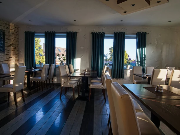 DSC07138_19La Terrazza.jpg Heller Gastraum mit dunklen Holztischen, weißen Stühlen und blau-weißen Vorhängen an den Fenstern.