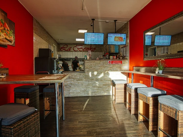 DSC07117_12la-benko.jpg Innenraum der Pizzeria mit roten Wänden, Rattan-Hockern und Tresenbereich im Hintergrund.