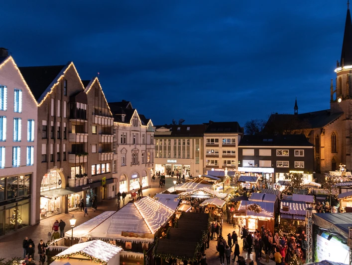 Impression Gütersloher Weihnachtsmarkt Ein Markt mit festlich beleuchteten Ständen auf einem Platz, umgeben von historischen Gebäuden bei Nacht.