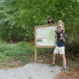 Auf der Familienvariante des Paderborner Höhenwegs Eine Familie posiert vor einer Infotafel, umgeben von üppigem Wald, auf einem Wanderweg.