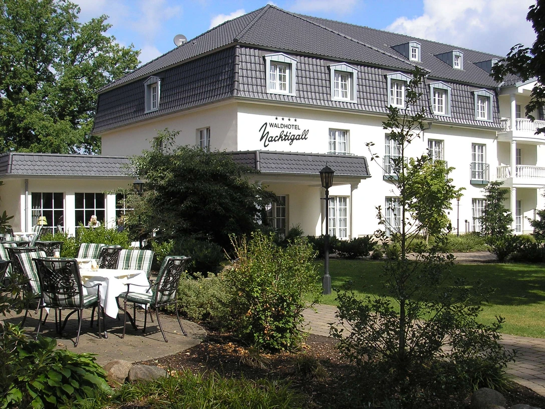 Waldhotel Nachtigall / Außenansicht Weiße Fassade eines dreistöckigen Hotels mit grauem Dach; begrünter Hof und Terrassenbestuhlung.