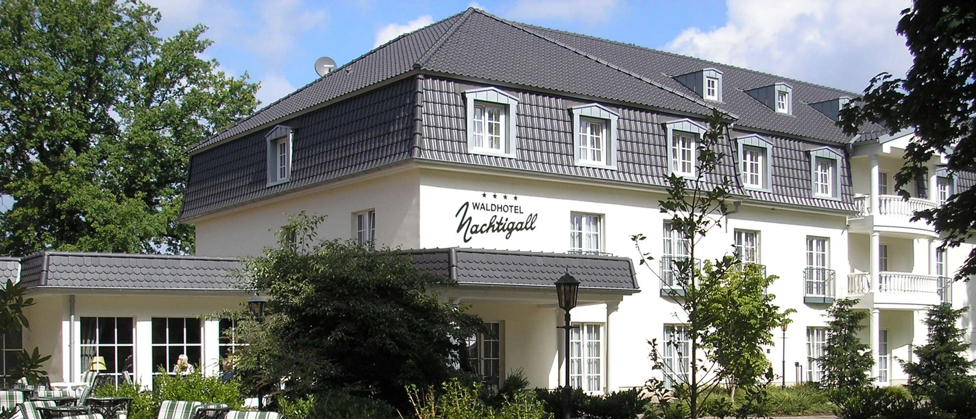 Waldhotel Nachtigall / Außenansicht Weiße Fassade eines dreistöckigen Hotels mit grauem Dach; begrünter Hof und Terrassenbestuhlung.