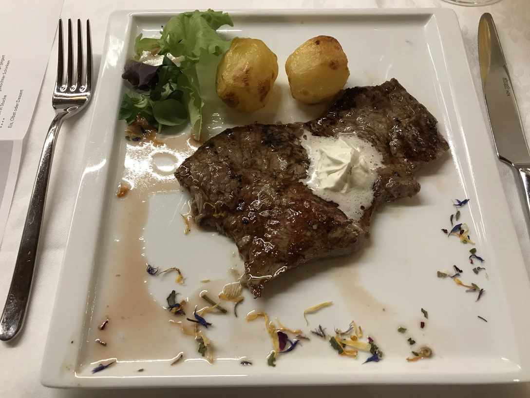 Steak Ein serviertes Steak auf einem weißen Teller, dekoriert mit essbaren Blüten, Salat und Kartoffeln.