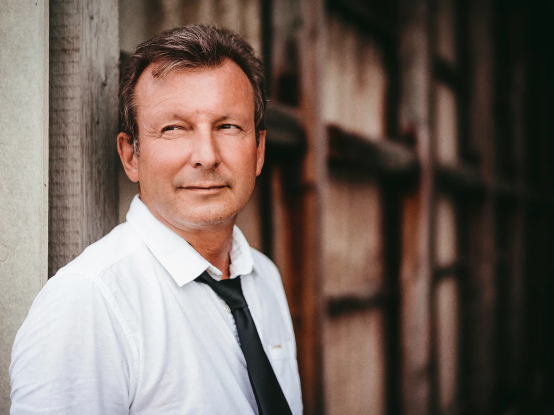 Frank-Suchland_1.jpg Ein Mann mittleren Alters in weißem Hemd und Krawatte, steht entspannt vor einer hölzernen Wand.