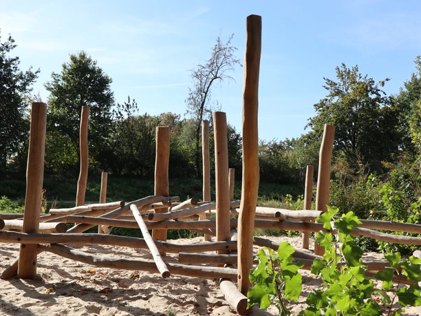Holzklettergerüst auf einem sandigen Spielplatz, umgeben von Bäumen unter klarem Himmel.