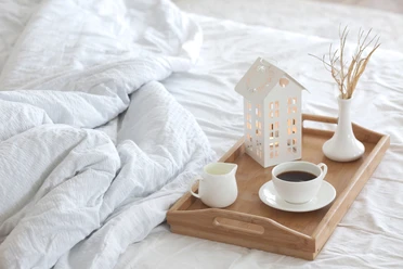 Ein Frühstückstablett mit Kaffee und Milch steht auf einem weißen Bett neben einer Deko-Laterne.