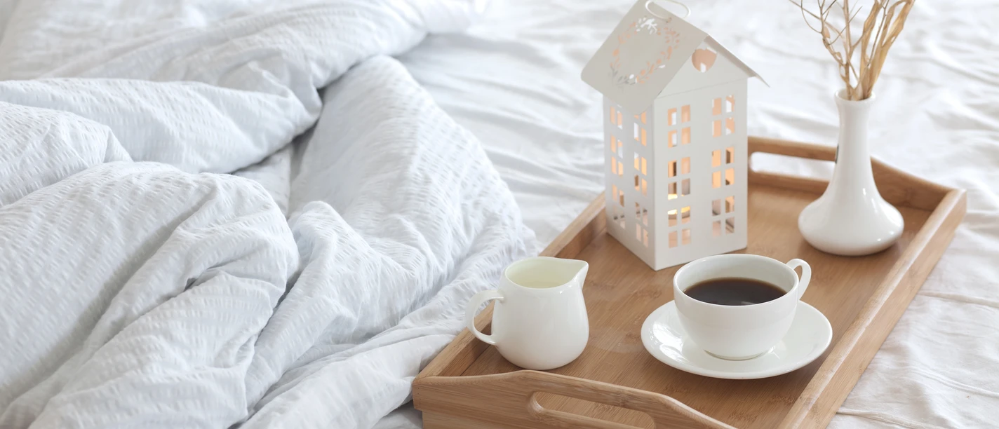 Ein Frühstückstablett mit Kaffee und Milch steht auf einem weißen Bett neben einer Deko-Laterne.