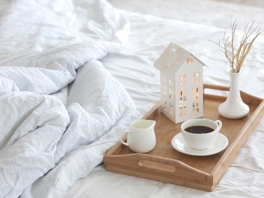 Musterfoto Übernachtung Ein Frühstückstablett mit Kaffee und Milch steht auf einem weißen Bett neben einer Deko-Laterne.