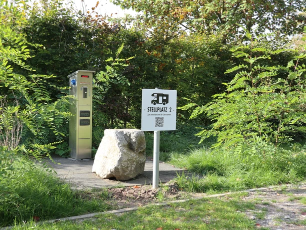 Versorgungsstation Stellplatz für Wohnmobile mit elektrischem Anschluss, umgeben von grünen Büschen und Bäumen.