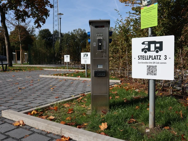 Stellplätze mit digitaler Anmeldung Ein Wohnmobilstellplatz im Herbst mit QR-Code-Schild und asphaltierter Stellfläche unter blauem Himmel.