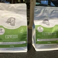 Neanderthaler Kaffee aus der Kult-Kaffee Rösterei in Heiligenhaus Zwei Kaffeebeutel aus der Kult-Kaffee Rösterei, links Espresso, rechts Filterkaffee, vor Regal.