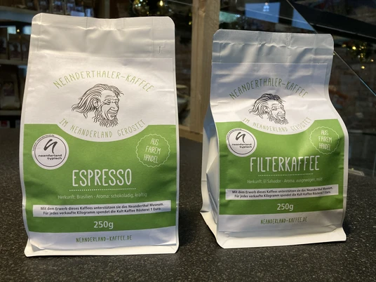 Neanderthaler Kaffee aus der Kult-Kaffee Rösterei in Heiligenhaus Zwei Kaffeebeutel aus der Kult-Kaffee Rösterei, links Espresso, rechts Filterkaffee, vor Regal.