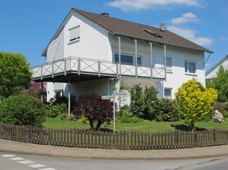 Modernes zweistöckiges Einfamilienhaus mit Balkon und gepflegtem Garten an einer Straßenecke.