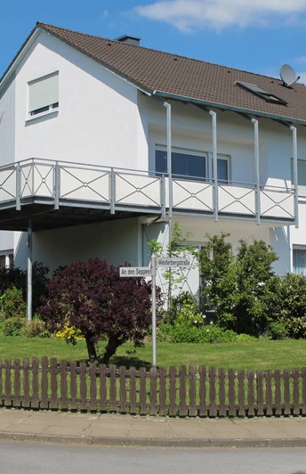 Modernes zweistöckiges Einfamilienhaus mit Balkon und gepflegtem Garten an einer Straßenecke.