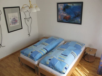 Zwei Einzelbetten mit blauer Bettwäsche in einem hellen Raum mit Holzboden und moderner Kunst an den Wänden.