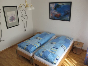 Zwei Einzelbetten mit blauer Bettwäsche in einem hellen Raum mit Holzboden und moderner Kunst an den Wänden.